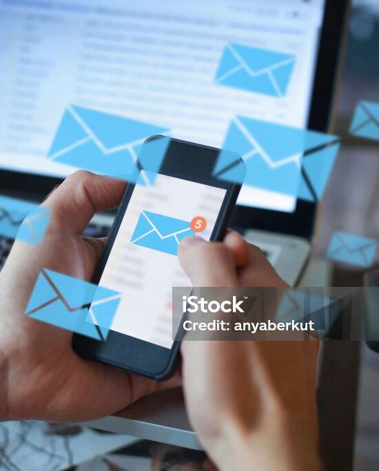 Person hält ein Smartphone mit einer neuen E-Mail-Benachrichtigung, während ein Laptop im Hintergrund offene E-Mails anzeigt – Symbol für digitale Kommunikation und E-Mail-Sicherheit.
